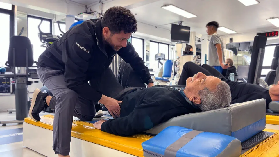 Kinesiólogo asistiendo a un paciente adulto mayor recostado en camilla durante sesión de kinesiología en KinesicWork