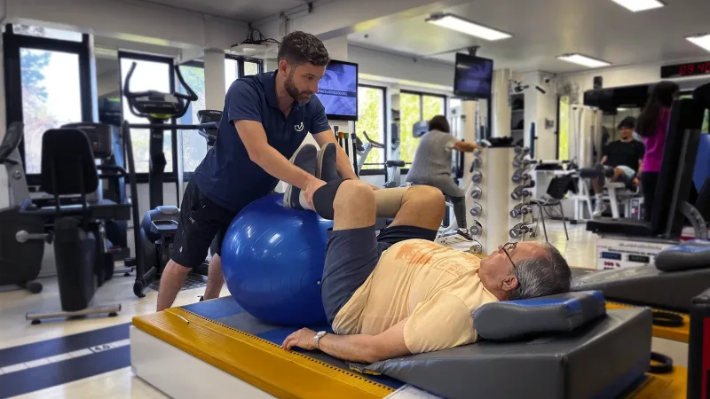 Kinesiólogo asistiendo a paciente adulto mayor recostado en colchoneta mientras realiza ejercicio con balón terapéutico en KinesicWork